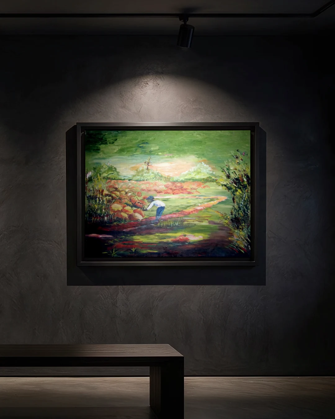 A painting in a thin black frame on a dark plaster wall under a warm spotlight: a green landscape: a figure in a white shirt and blue trousers bends over beside a path, among red and ochre rocks, under a pale glowing sky, with dense foliage at the right edge.