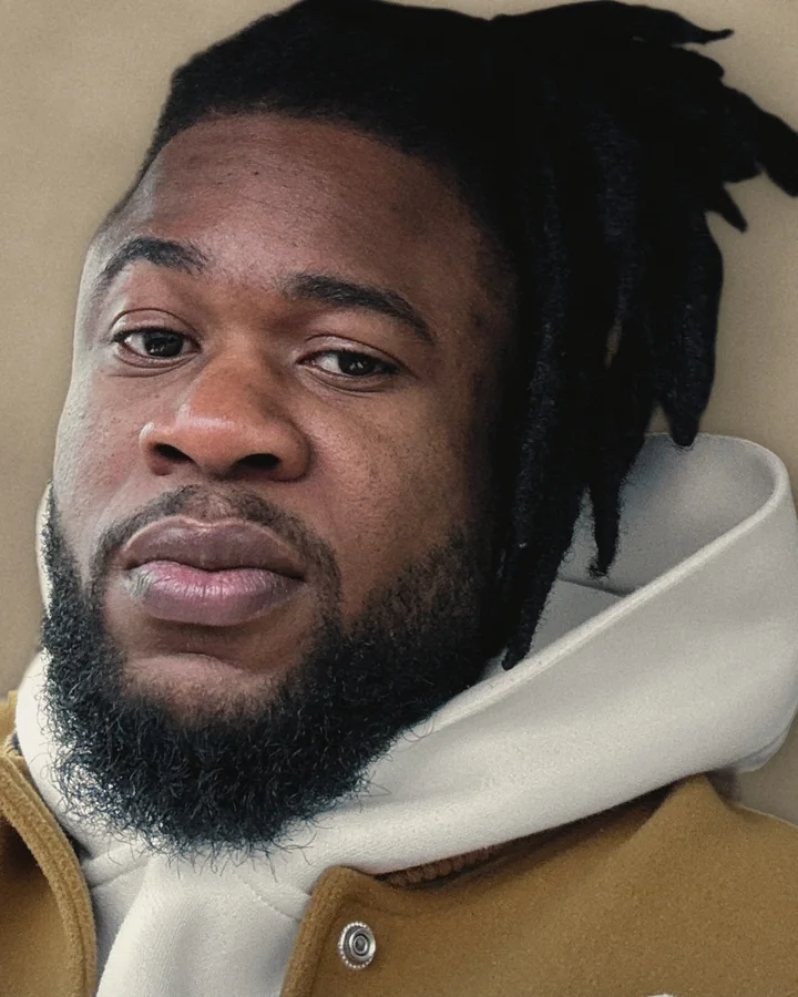 David Akintade, close up, head and shoulders. He has locs and a full beard, wears an ivory hoodie under a mustard jacket, and looks straight at the camera.